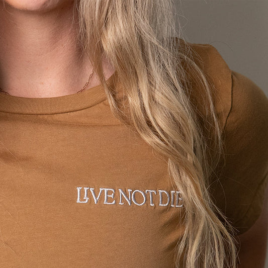 Brown t-shirt with 'LIVE NOT DIE' text worn by a person with blonde hair.