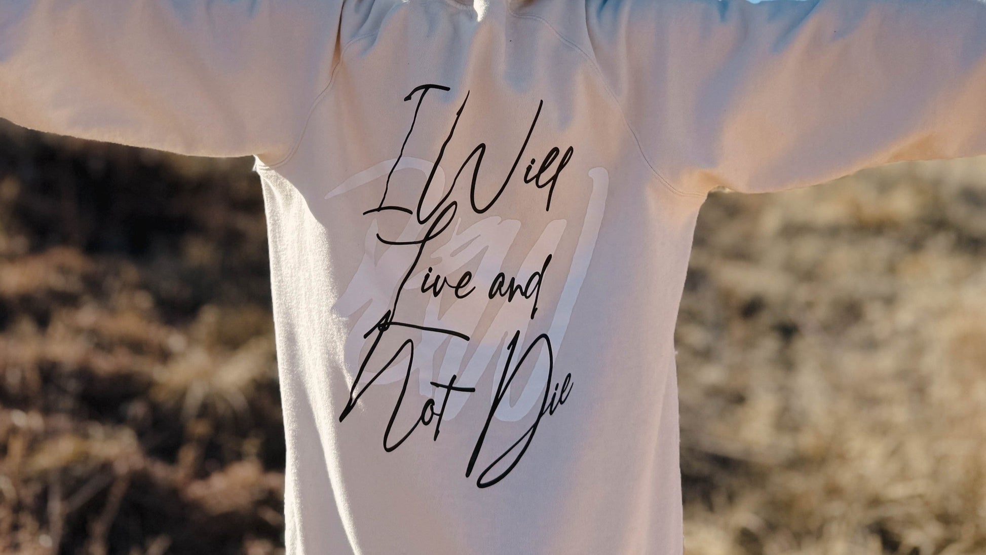 A women in a field wearing a LIVE NOT DIE branded crewneck and a brown beanie