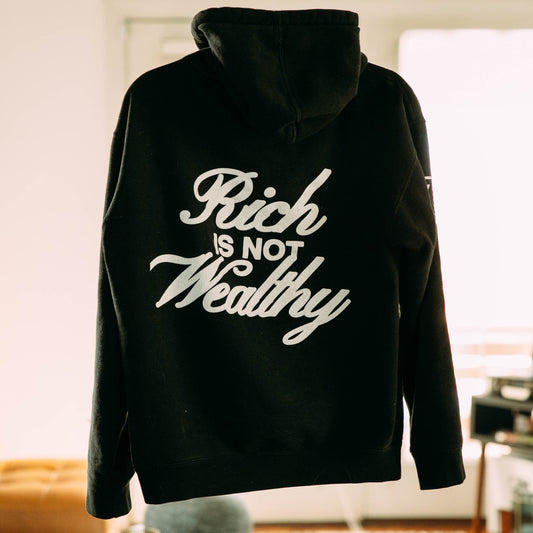 The black Rich Is Not Wealthy hoodie in a living room