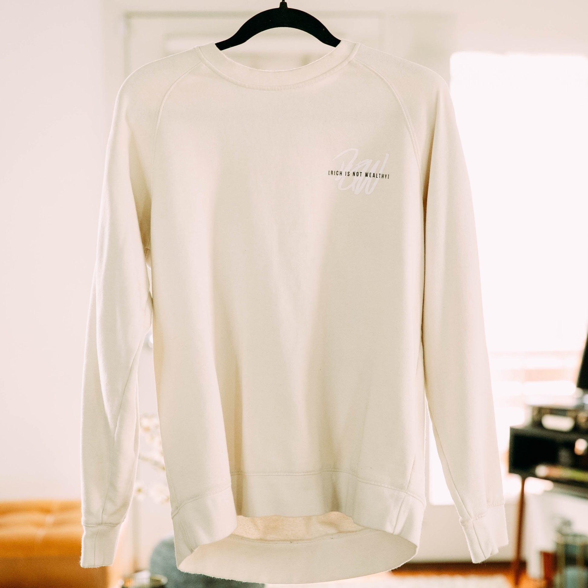 Front of the I Will Live Not Sweatshirt in a living room with rich is not wealthy logo